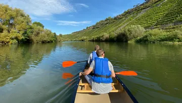 Two people canoeing on a river with vineyard hills.