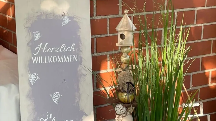 Willkommensschild mit Pflanzen und Vogelhaus an einer Backsteinmauer.