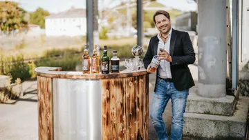 Man standing by outdoor wooden bar with whiskey bottles.