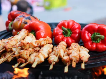 Hähnchen-Spieße grillen neben roten Paprika auf Grill.