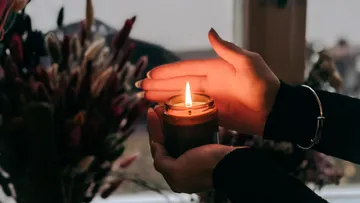 Hände, die eine brennende Kerze in der Nähe von Blumen halten.