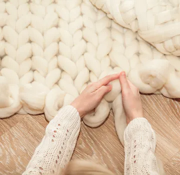 Hands knitting chunky wool blanket on floor.