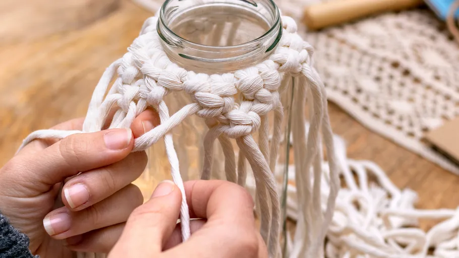 Hands crafting macramé jar holder with knots.