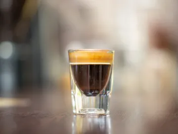 Ein Glas Espresso steht auf einem holzigen Tisch.