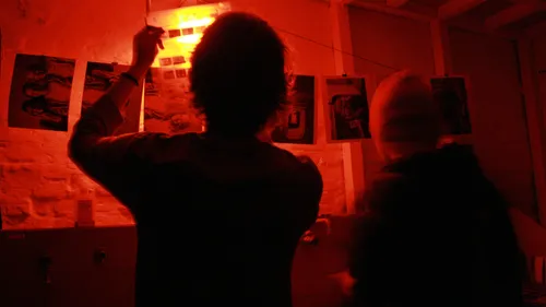 Zwei Personen entwickeln Fotos in einer rot beleuchteten Dunkelkammer.