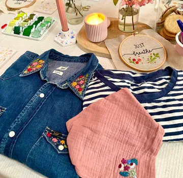 Denim shirt, striped t-shirt, and pink cloth with embroidery on table with candle and embroidery hoop.