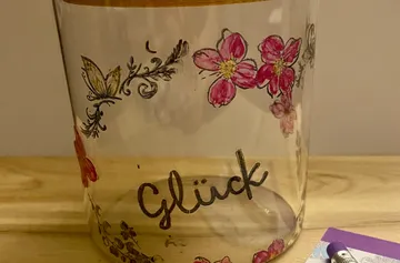 Decorated jar with flowers, text 'Glück', on table.