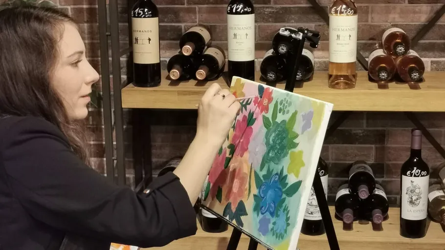 Frau malt bunte Blumen auf Leinwand, Weinflaschen dahinter.