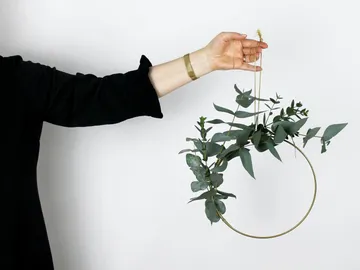 Hand holds plants in a golden ring, against a neutral background.