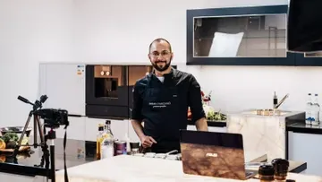 Chef in kitchen with laptop and camera setup.