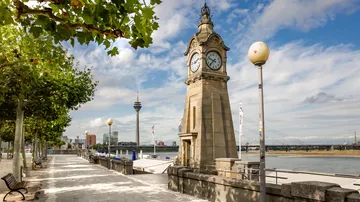 Clock tower on riverside promenade with trees.