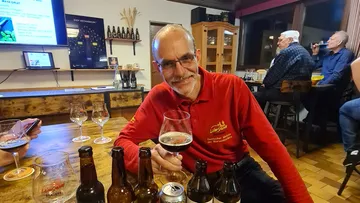 Man in red shirt with beer glass smiles in brewery.