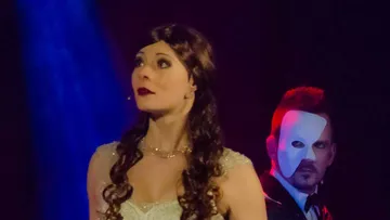 Frau singt mit maskiertem Mann auf der Bühne.