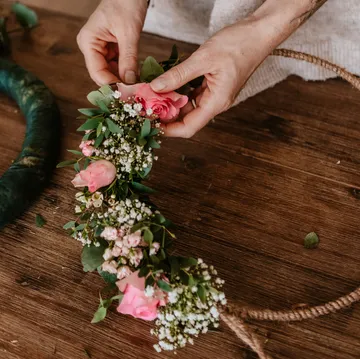 Hände beim Arrangieren von Blumen auf einem Kranzrahmen.