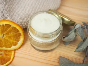 A glass cream lies next to a dried orange and herbs.