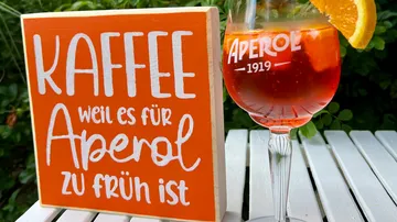 Auf dem Schild steht „KAFFEE weil es für Aperol zu früh ist“ neben dem Aperolglas auf dem Tisch.