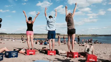 Drei Personen stehen auf Kisten am Strand.