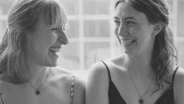 Two women smiling indoors, wearing necklaces.