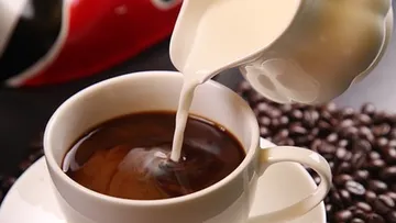 Milch in eine Kaffeetasse gießen, umgeben von Bohnen.