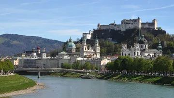 Fortress and cityscape beside a river, with mountains.