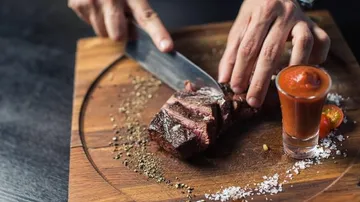 Person slicing steak on wooden board with sauce.