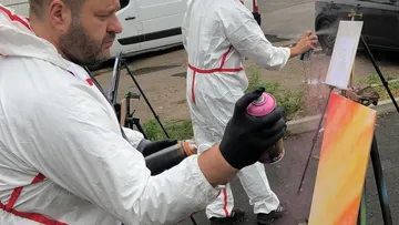 Two artists spray paint canvases outdoors.