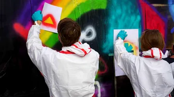 Two people spray stencil on graffiti wall.