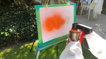 Person spray-painting 'VASILY' on stencil outdoors.