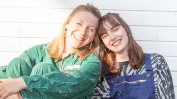 Zwei Frauen lächeln draußen in Freizeitkleidung.