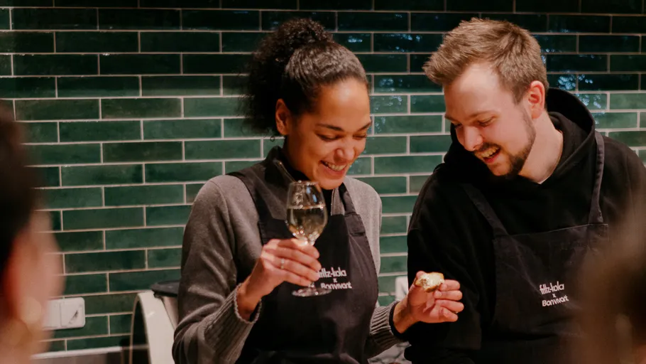 Two people wearing aprons, holding wine and food in kitchen.
