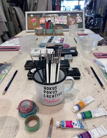 Art supplies on table with paintbrushes in a mug.