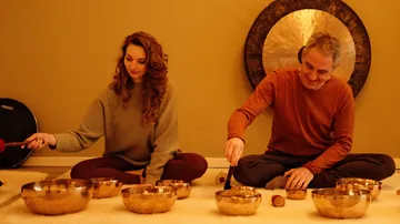 Two people play singing bowls indoors.