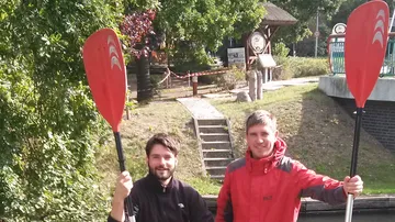 Zwei Leute, die draußen Paddel halten und lächeln.
