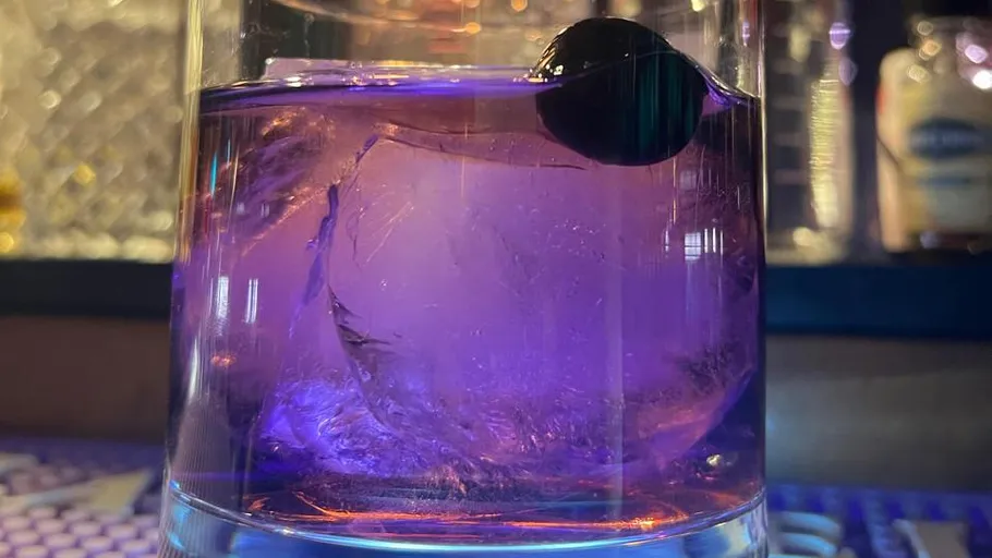 Purple cocktail with large ice cube and cherry.