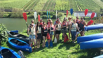 Group with paddles by river and canoes, vineyard background.