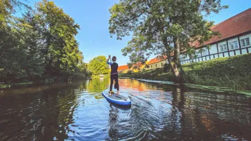 Person beim Paddeln auf einem ruhigen Fluss in der Nähe von Bäumen und Häusern.