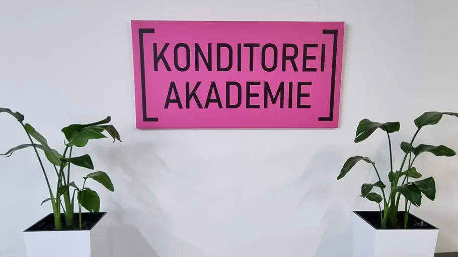 Rosafarbenes Schild mit der Aufschrift „Konditorei Akademie“ an der Wand, flankiert von Topfpflanzen.