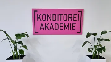 Rosafarbenes Schild mit der Aufschrift „KONDITOREI AKADEMIE“ zwischen Pflanzen.