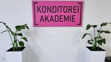 Rosafarbenes Schild mit der Aufschrift „Konditorei Akademie“ zwischen zwei Pflanzen.