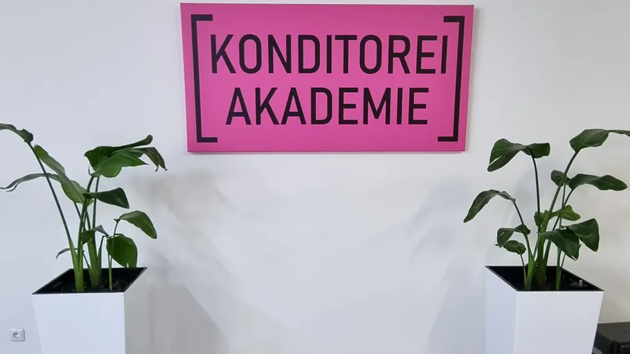 Rosafarbenes Schild mit 'Konditorei Akademie' zwischen zwei Pflanzen.