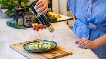Person in blue dress pouring olive oil on cheese plate.