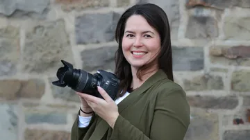 Woman holding camera, standing by brick wall.