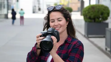 Woman holding camera, smiling outdoors.