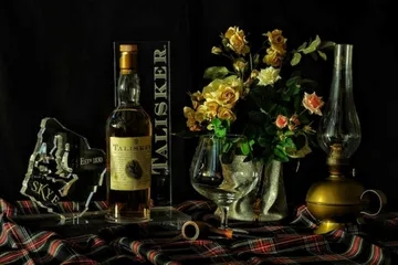 Whiskey bottle, glass, flowers, lamp on cloth.