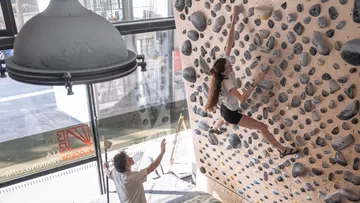 Frau klettert auf eine Indoor-Boulderwand; eine Person entdeckt sie.