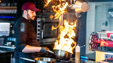 Koch hält Pfanne mit großen Flammen in der Küche.