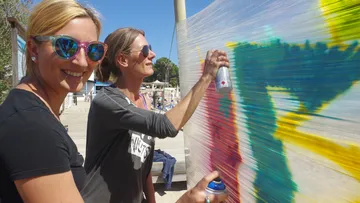 Two women spray paint on plastic outdoors.