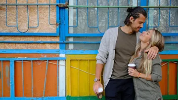 Couple smiles with coffee by colorful wall.