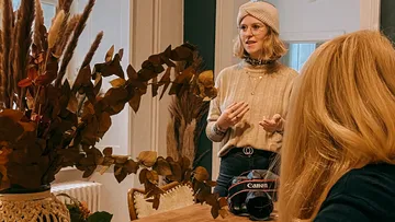 Person talking near a vase of tall dried plants.