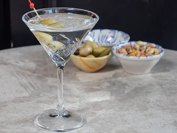 Martini glass with olives is on a table with snacks in the background.
